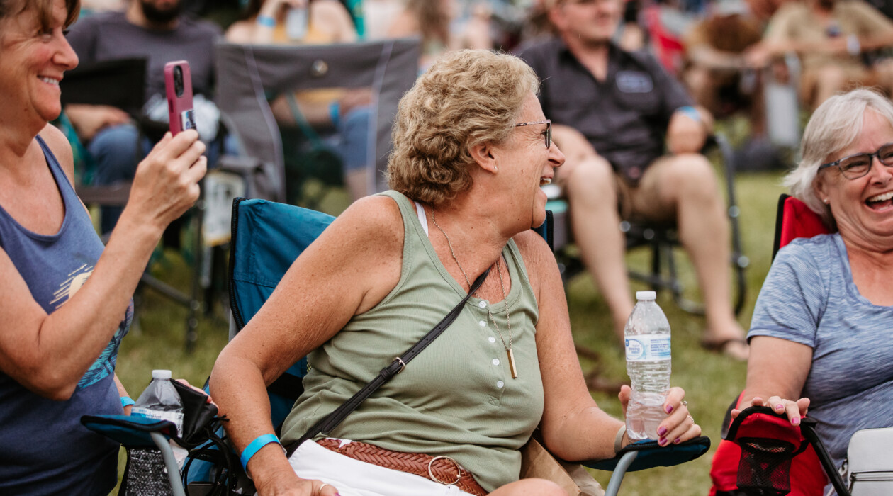 One woman takes a photo of two others laughing, in the front row of the Fieldstone Music Festival.